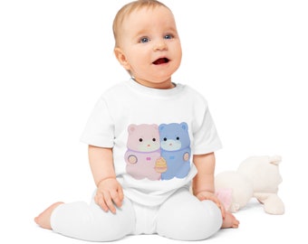 May include: A baby wearing a white onesie with a pink and blue bear design. The bears are holding a small yellow cupcake. The baby is sitting on a white background.