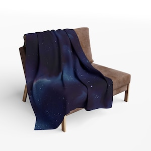 Peut inclure: Une couverture en jetée bleu foncé avec un imprimé galaxie représentant des étoiles blanches et une nébuleuse. La couverture est drapée sur un fauteuil marron.