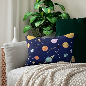 May include: A navy blue throw pillow with a pattern of planets, stars, and swirls in yellow, orange, pink, and blue. The pillow is on a white couch with a white blanket.
