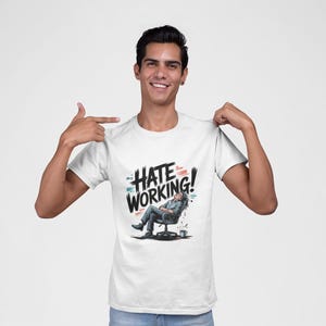 Può includere: T-shirt bianca con la scritta "HATE WORKING!" in un carattere audace e stilizzato. La grafica presenta una persona che si rilassa su una sedia da ufficio. La persona nell'immagine indica la maglietta.