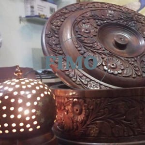 Può includere: Una ciotola da portata in legno marrone scuro, intagliata in modo intricato, con un coperchio abbinato. Il coperchio e la ciotola presentano motivi floreali e a foglie. In primo piano, una lampada rotonda in metallo marrone più piccola con ritagli a forma di stella.