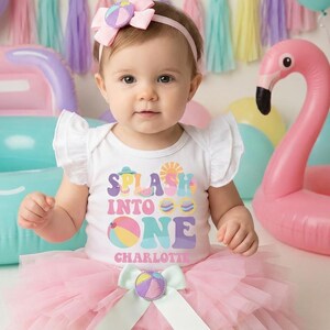 以下が含まれることがあります： 「Splash Into One Charlotte」の文字が入った白いボディスーツとピンクのチュチュを着た赤ちゃん。背景には「Happy Birthday Charlotte」と書かれたバナーとピンクのインフレータブルフラミンゴがあります。