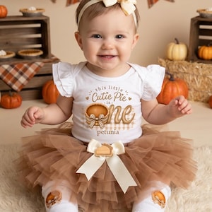 Puede incluir: Un body blanco de manga corta con volantes que incluye el texto this little Cutie Pie is one y el nombre Petunia, combinado con un tutú de tul marrón, leggings blancos con gráficos de tartas, una diadema color crema y zapatos dorados.
