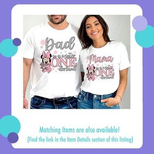 May include: White t-shirts with matching designs. One shirt says "Dad" and the other says "Mama" in gray script, with "in a winter ONE-derland" below. Each shirt features a Minnie Mouse graphic in pink. The shirts are worn by two people.