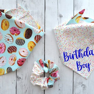 May include: Two dog bandanas and a scrunchie. One bandana features a light blue background with donut illustrations. The other bandana has a white background with colorful sprinkles and the text "Birthday Boy". The scrunchie has a donut and sprinkle pattern.