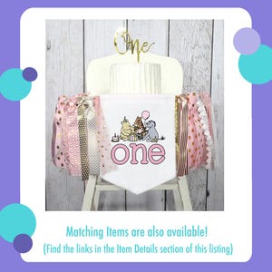 Puede incluir: Una tarta blanca con un adorno dorado de "One", decorada con una pancarta con los personajes de Winnie the Pooh y la palabra "one" en rosa. Cintas rosas y doradas adornan los lados, creando un aspecto festivo.