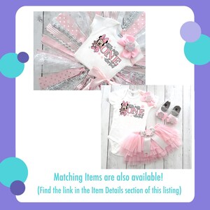 May include: A white baby bodysuit with a Minnie Mouse graphic and the word "ONE" in pink. The outfit includes a pink tutu, a matching bow, and baby shoes. The set is designed for a first birthday celebration.