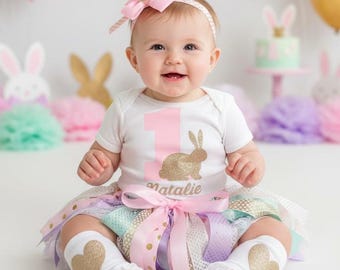 Puede incluir: Un bebé con un body blanco con el número 1 rosa y un conejo dorado con purpurina. El bebé lleva un tutú multicolor, zapatos con detalles dorados y calcetines blancos con corazones dorados. Un lazo rosa adorna la cabeza del bebé.