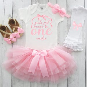 May include: A baby girl's first birthday outfit set. The set includes a white onesie with pink text that reads "girls just wanna be one amelia", a pink tutu skirt, a pink bow headband, white leg warmers with pink bows, and gold shoes with pink bows.