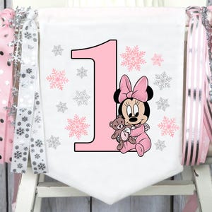 May include: A white banner with a large pink number 1, pink snowflakes, and a Minnie Mouse graphic holding a teddy bear. The banner is decorated with pink and silver ribbons.