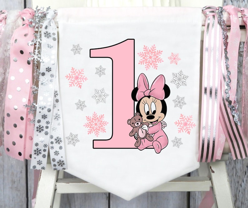 May include: A white banner with a large pink number 1, pink snowflakes, and a Minnie Mouse graphic holding a teddy bear. The banner is decorated with pink and silver ribbons.