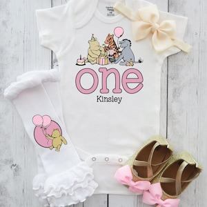 Puede incluir: Conjunto de beb&eacute; blanco con estampado rosa "One" y el nombre "Kinsley". El conjunto incluye un body, una diadema a juego, calentadores de piernas con volantes y zapatos dorados con purpurina y lazos rosas.