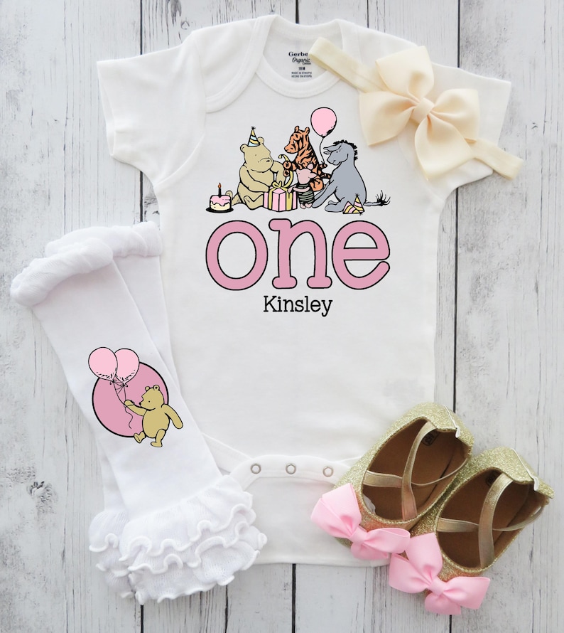 Puede incluir: Conjunto de beb&eacute; blanco con estampado rosa "One" y el nombre "Kinsley". El conjunto incluye un body, una diadema a juego, calentadores de piernas con volantes y zapatos dorados con purpurina y lazos rosas.