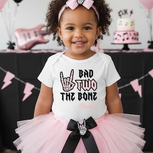 May include: A young child wearing a white onesie with the text "Bad Two The Bone" and a pink tutu. The outfit is accessorized with a pink bow headband and a black bow with a rock-and-roll hand symbol. Balloons and a cake are in the background.
