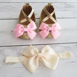 Puede incluir: Un conjunto de zapatos de beb&eacute; y una diadema. Los zapatos son dorados con lazos rosas y correas cruzadas. La diadema tiene un lazo color crema. Los art&iacute;culos est&aacute;n sobre una superficie clara con vetas de madera.