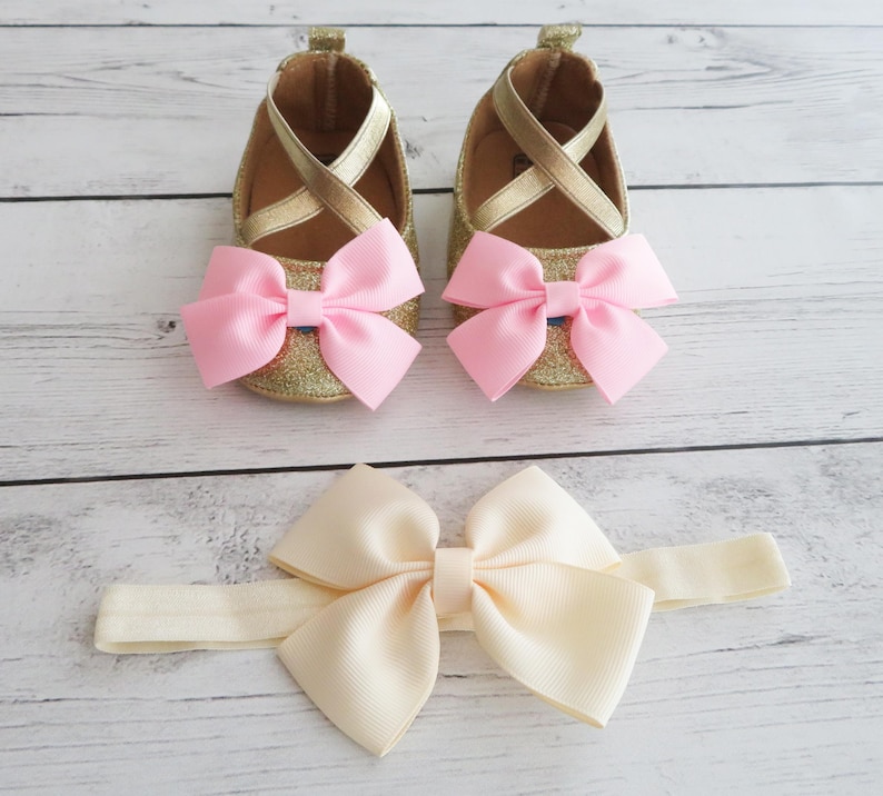 Puede incluir: Un conjunto de zapatos de beb&eacute; y una diadema. Los zapatos son dorados con lazos rosas y correas cruzadas. La diadema tiene un lazo color crema. Los art&iacute;culos est&aacute;n sobre una superficie clara con vetas de madera.