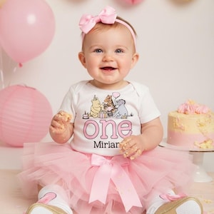 Puede incluir: Un beb&eacute; con un body blanco con un dise&ntilde;o de Winnie the Pooh y la palabra "One" en rosa, combinado con un tut&uacute; y un lazo rosa. El beb&eacute; sonr&iacute;e, rodeado de globos rosas, decoraciones de papel y una tarta.