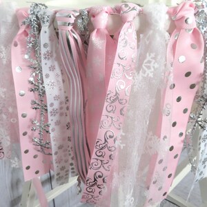 May include: A close-up of a decorative garland featuring pink and white ribbons with silver accents. The ribbons have snowflake and polka dot patterns. Silver snowflake ornaments are also included.