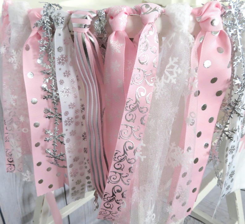 May include: A close-up of a decorative garland featuring pink and white ribbons with silver accents. The ribbons have snowflake and polka dot patterns. Silver snowflake ornaments are also included.
