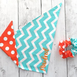 May include: A set of pet accessories, including a bandana and scrunchie. The bandana features a chevron pattern in turquoise and white, and a polka dot pattern in orange and white. The name "Manfred" is printed on the bandana. The scrunchie has matching patterns.