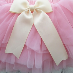 Puede incluir: Falda de tul rosa con m&uacute;ltiples capas, adornada con un gran lazo color crema. El lazo tiene largas cintas sueltas. La falda parece suave y esponjosa, adecuada para un atuendo infantil.
