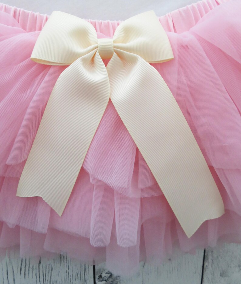 Puede incluir: Falda de tul rosa con m&uacute;ltiples capas, adornada con un gran lazo color crema. El lazo tiene largas cintas sueltas. La falda parece suave y esponjosa, adecuada para un atuendo infantil.