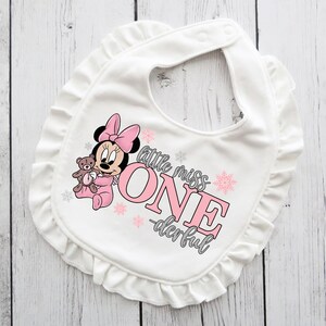 May include: White baby bib with a ruffled edge. The bib features a Minnie Mouse graphic in pink, holding a teddy bear. Text on the bib reads "little miss ONE -derful" in pink and gray.