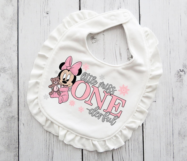 May include: White baby bib with a ruffled edge. The bib features a Minnie Mouse graphic in pink, holding a teddy bear. Text on the bib reads "little miss ONE -derful" in pink and gray.