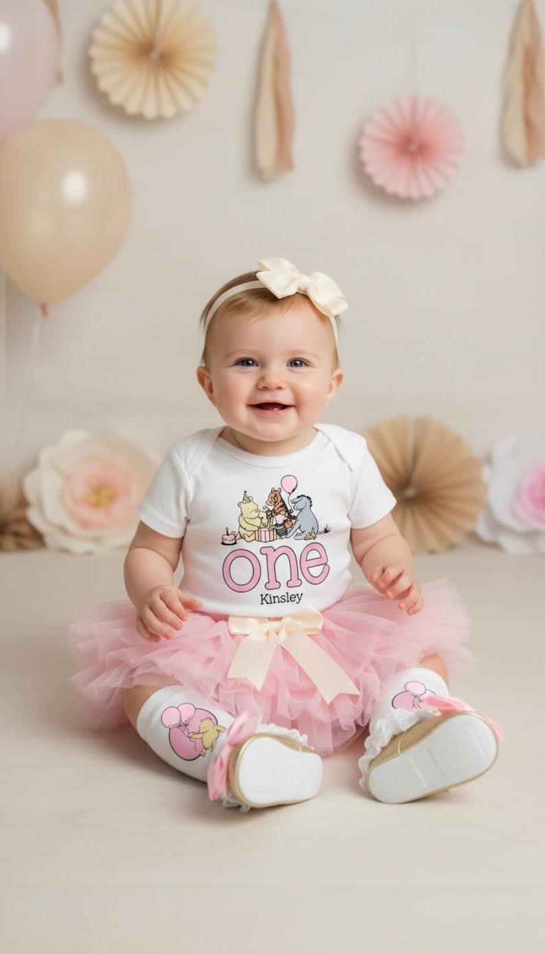 Puede incluir: Un beb&eacute; sonriente con un body blanco con un gr&aacute;fico de Winnie the Pooh y la palabra "One" en rosa. El beb&eacute; tambi&eacute;n lleva un tut&uacute; rosa, calcetines, una diadema con un lazo y zapatos blancos con suelas doradas. El nombre "Kinsley" est&aacute; debajo de la palabra "One".