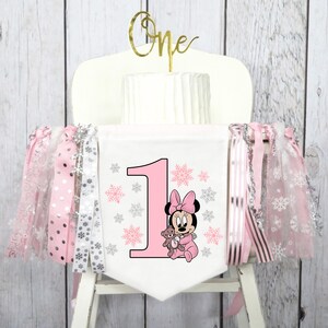 May include: A white high chair banner with a pink number one, Minnie Mouse graphic, and snowflake accents. The banner is decorated with pink and silver ribbons. A gold "One" cake topper sits atop a white cake.