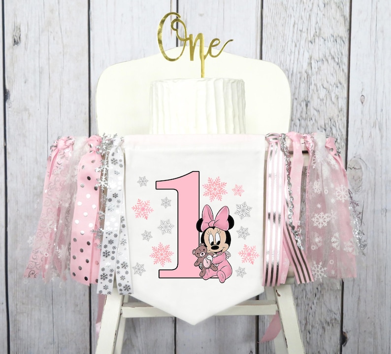 May include: A white high chair banner with a pink number one, Minnie Mouse graphic, and snowflake accents. The banner is decorated with pink and silver ribbons. A gold "One" cake topper sits atop a white cake.