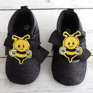 Puede incluir: Par de zapatos de bebé negros con purpurina, con una aplicación de abeja amarilla y negra y un lazo negro. Los zapatos son de talla 3.