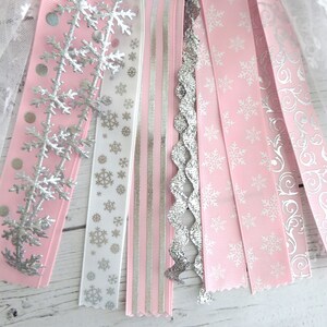 May include: A collection of decorative ribbons in shades of pink, white, and silver. The ribbons feature snowflake and swirl patterns, along with silver glitter and polka dots. The ribbons are arranged in a fan shape, suggesting a festive or holiday theme.