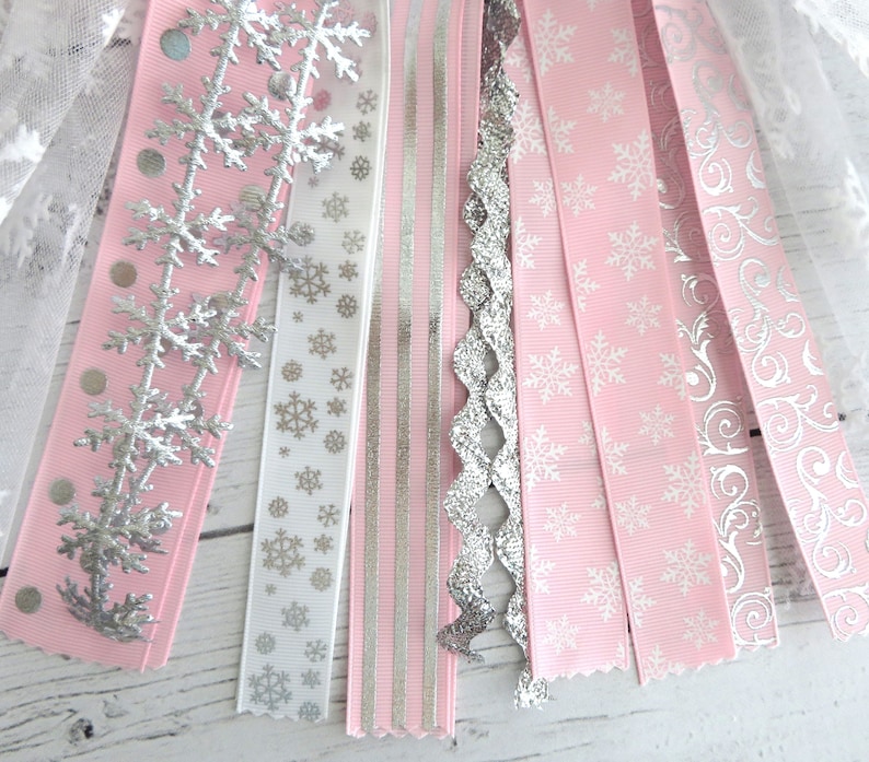 May include: A collection of decorative ribbons in shades of pink, white, and silver. The ribbons feature snowflake and swirl patterns, along with silver glitter and polka dots. The ribbons are arranged in a fan shape, suggesting a festive or holiday theme.
