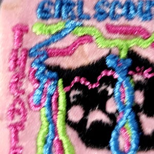 May include: Embroidered patch with the words "GIRL SCOUT" in blue thread. The patch features a black paw print and colorful, wavy lines in pink, blue, and green. The background is a light pink.
