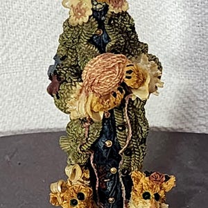 Pode incluir: Estatueta detalhada de um urso de peluche com um casaco verde e um chapéu com rosas vermelhas. O urso está a segurar um novelo de lã e é ladeado por duas figuras de ursos mais pequenos. A estatueta é pintada em tons suaves.