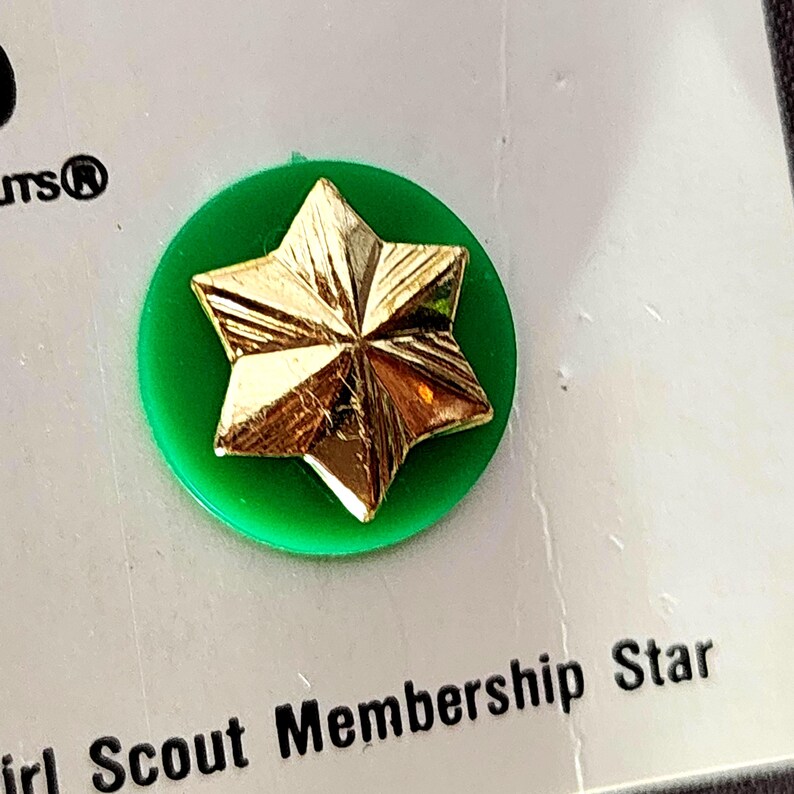 May include: A gold-colored star pin on a green circular base. The pin is a Girl Scout Membership Star. The text "Girl Scout Membership Star" is visible below the pin.