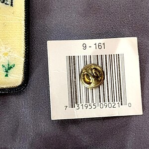 May include: A small, round, gold-colored pin with a decorative design. The pin is attached to a white tag with a barcode and the numbers "9-161". The background is a dark gray fabric.
