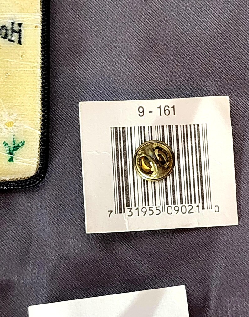May include: A small, round, gold-colored pin with a decorative design. The pin is attached to a white tag with a barcode and the numbers "9-161". The background is a dark gray fabric.