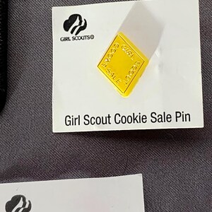 May include: A yellow Girl Scout Cookie Sale pin on a white card. The pin is square with rounded corners and the words "Girl Scout Cookie Sale" are printed on it. The card has the Girl Scouts logo and the text "Girl Scout Cookie Sale Pin".
