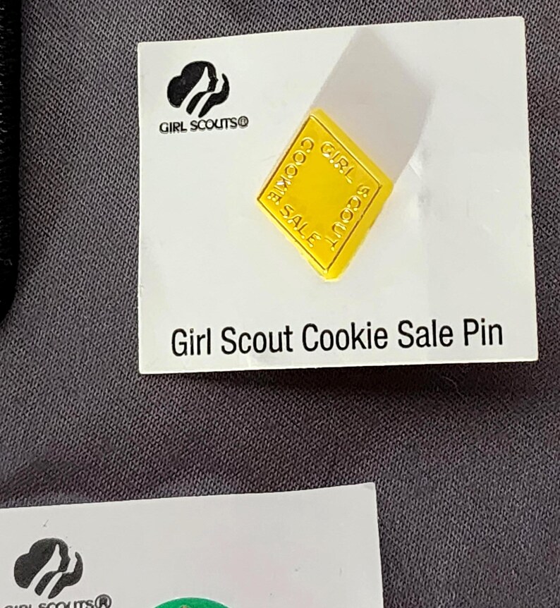 May include: A yellow Girl Scout Cookie Sale pin on a white card. The pin is square with rounded corners and the words "Girl Scout Cookie Sale" are printed on it. The card has the Girl Scouts logo and the text "Girl Scout Cookie Sale Pin".