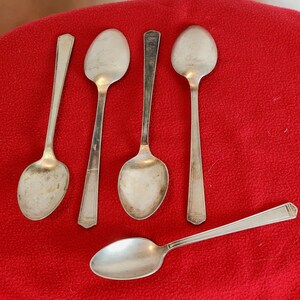 Puede incluir: Cinco cucharas de plata de diferentes diseños dispuestas sobre una superficie roja. Las cucharas tienen cuencos ovalados y mangos largos. Algunos mangos tienen un diseño geométrico en el extremo. Las cucharas son probablemente vintage o antiguas.