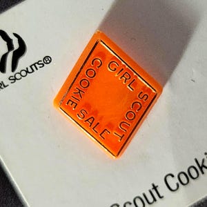 May include: An orange, diamond-shaped Girl Scout Cookie Sale pin. The pin has the words "COOKIE SALE" and "GIRL SCOUT" in a gold color around the edges. The pin is on a white surface with the Girl Scouts logo.