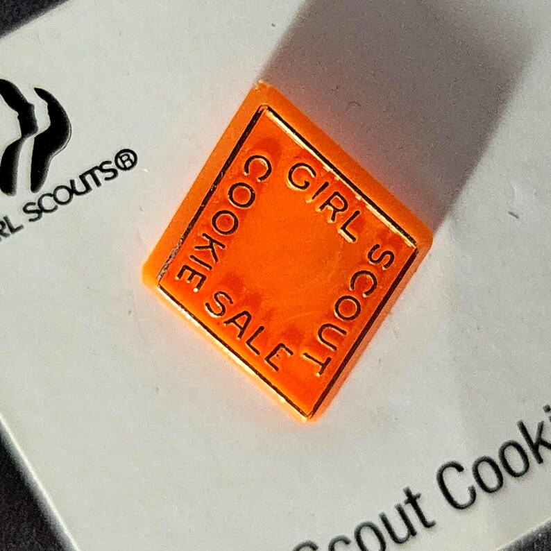May include: An orange, diamond-shaped Girl Scout Cookie Sale pin. The pin has the words "COOKIE SALE" and "GIRL SCOUT" in a gold color around the edges. The pin is on a white surface with the Girl Scouts logo.