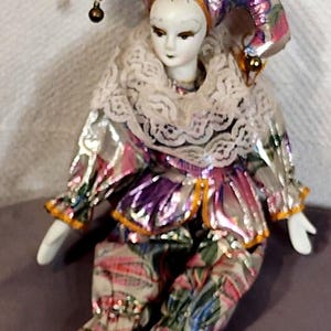 Peut inclure: Une poupée bouffon en porcelaine au visage peint, vêtue d'une tenue en tissu métallique multicolore avec des motifs floraux roses, violets et verts. La poupée porte un col en dentelle blanche et un chapeau à trois pointes avec des grelots dorés.