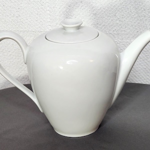 May include: A white ceramic teapot with a curved handle and a long, slender spout. The teapot has a matching lid with a small knob. The design is simple and elegant, suitable for serving tea.