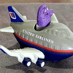 May include: A toy airplane with the United Airlines logo, featuring a purple character in the cockpit. The plane is gray, blue, and red, with a white secondary plane attached below.