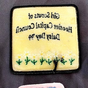 May include: A square, yellow patch with black trim featuring text and a daisy design. The text reads "Girl Scouts of Houston Capital Council Dairy Day". The bottom of the patch has a row of white daisies with yellow centers and green stems.