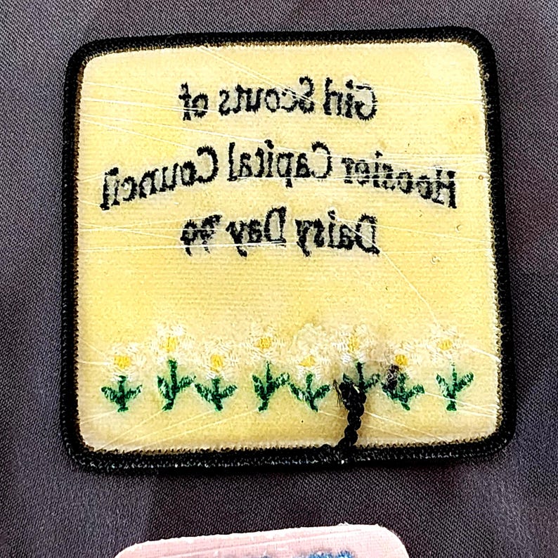 May include: A square, yellow patch with black trim featuring text and a daisy design. The text reads "Girl Scouts of Houston Capital Council Dairy Day". The bottom of the patch has a row of white daisies with yellow centers and green stems.