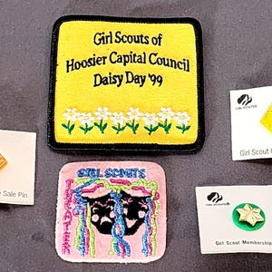 以下が含まれることがあります： ガールスカウトの記念品コレクション。パッチとピンが含まれています。黒い縁取りのある黄色のパッチには「Girl Scouts of Hoosier Capital Council Daisy Day '99」と書かれています。その他のアイテムには、ピンクのパッチとクッキー販売ピンが含まれます。